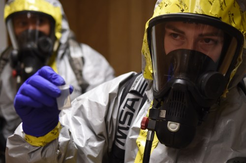 Two individuals in hazmat suits, one holding a small vial, both wearing masks and gloves.