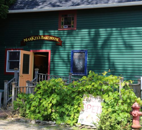 A green building with a sign reading "Markel's Bakehouse," featuring a welcoming entrance and lush greenery.