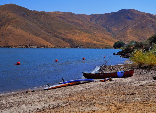 A serene lakeside scene with a canoe and paddleboards on the shore, surrounded by rolling hills and calm water.