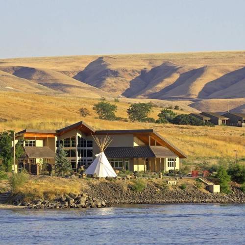 A modern building with a teepee by a river, surrounded by dry hills and greenery under a clear blue sky.