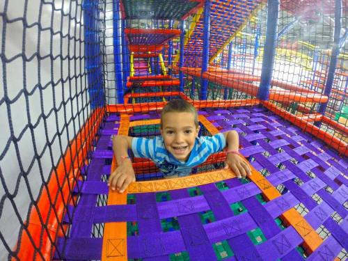 A smiling child peeks through colorful netting in a play structure filled with vibrant mats and tunnels.