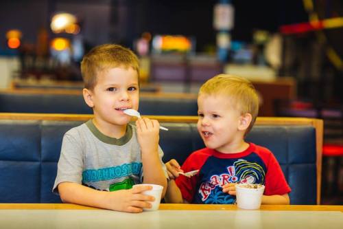 Two young boys sitting at a table, enjoying ice cream from cups and smiling at each other.