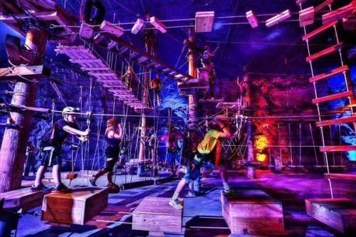 A vibrant indoor ropes course with climbers navigating obstacles and wooden platforms under colorful lighting.