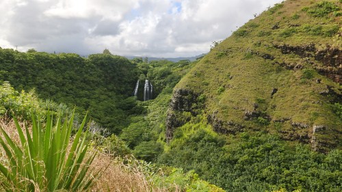 Lush green valley with a waterfall cascading down rocky cliffs under a cloudy sky.