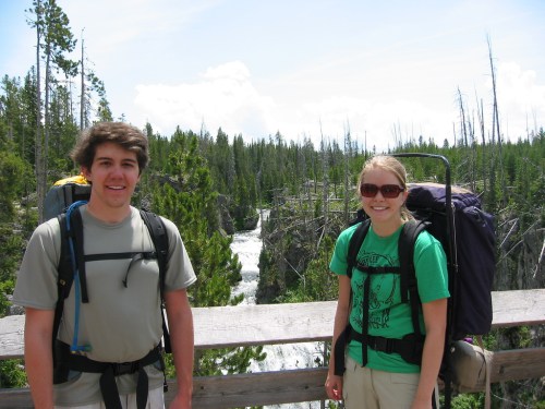Two hikers stand on a wooden overlook, smiling, with a waterfall and forested landscape in the background.