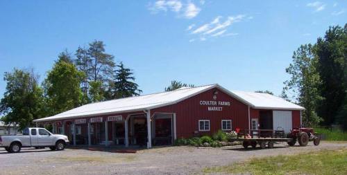 Red barn-style market building with a white roof, surrounded by trees and a gravel parking area.