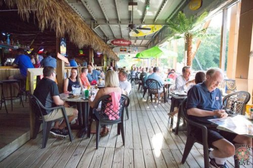 A lively outdoor restaurant scene with diners at tables under a thatched roof, surrounded by greenery and colorful decor.