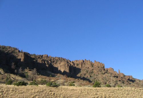 A rugged mountain range under a clear blue sky, with rocky formations and sparse vegetation in the foreground.