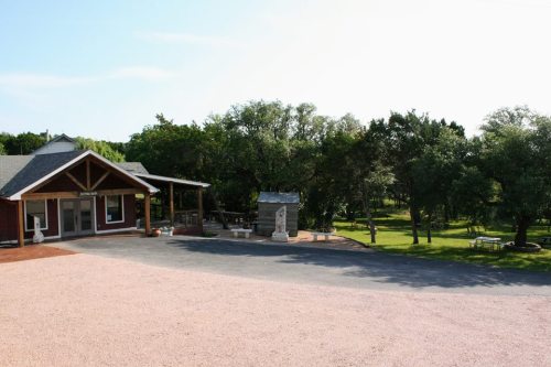 A rustic building surrounded by trees and a gravel path, with outdoor seating and a stone monument nearby.
