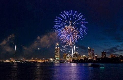 Colorful fireworks illuminate the night sky over a city skyline, reflecting on the water below.
