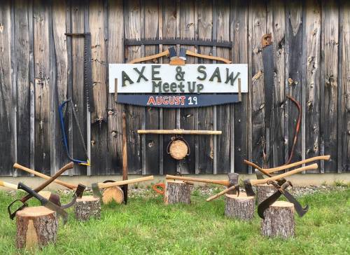 A rustic display featuring axes and saws arranged around wooden stumps, with a sign for an "Axe & Saw Meet Up" on August 19.