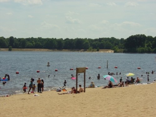 A sandy beach with people swimming and relaxing by a lake, colorful umbrellas dotting the shore under a sunny sky.