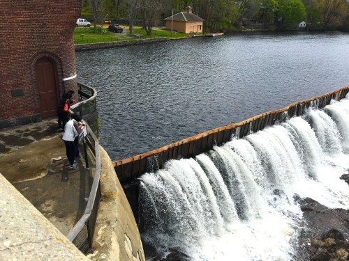 A view of a waterfall cascading over a dam, with people observing from a walkway nearby. Lush greenery surrounds the scene.