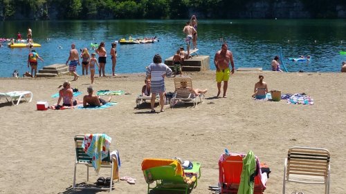 A busy beach scene with people sunbathing, swimming, and relaxing by a lake on a sunny day.