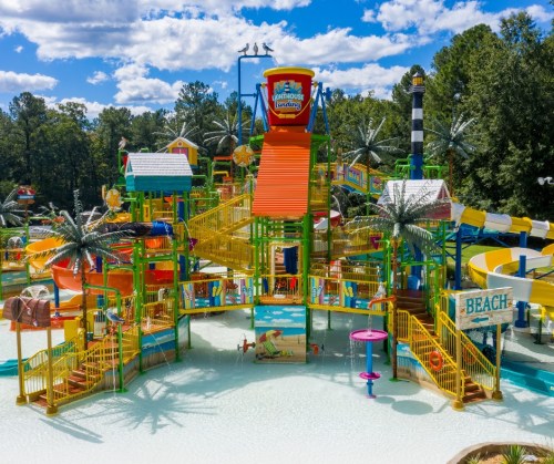 Colorful water park with slides, pools, and palm trees under a blue sky with clouds. Fun attractions for all ages.