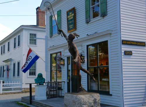 A bronze statue of a girl with a tennis racket stands outside a gallery with green shutters and an open sign.