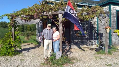 An elderly couple stands together outside a rustic building adorned with vines and a flag.