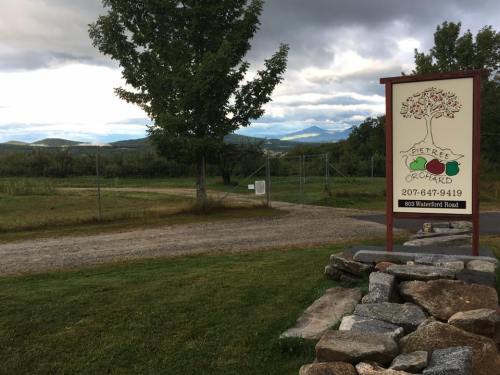 Sign for Tree Orchard with a scenic view of hills and cloudy sky in the background, surrounded by gravel path and trees.