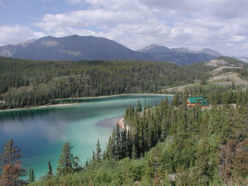 A serene lake surrounded by lush forests and mountains under a cloudy sky, with a cabin visible on the shore.