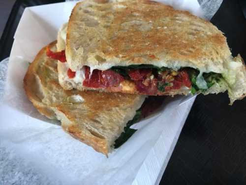 A toasted sandwich filled with greens, tomatoes, and cheese, served in a paper wrapper.