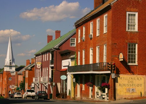 Historic brick buildings line a quaint street, with a church steeple visible in the background under a blue sky.
