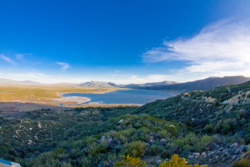 Panoramic view of a serene lake surrounded by rolling hills and mountains under a clear blue sky.