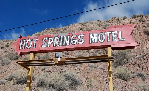 Sign for "Hot Springs Motel" against a rocky landscape with blue skies and scattered shrubs.