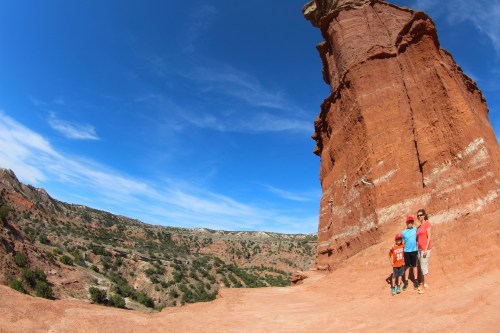 A family stands together in front of a tall red rock formation under a blue sky with scattered clouds.