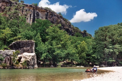 A serene river scene with a person relaxing on the shore, surrounded by lush greenery and rocky cliffs under a blue sky.