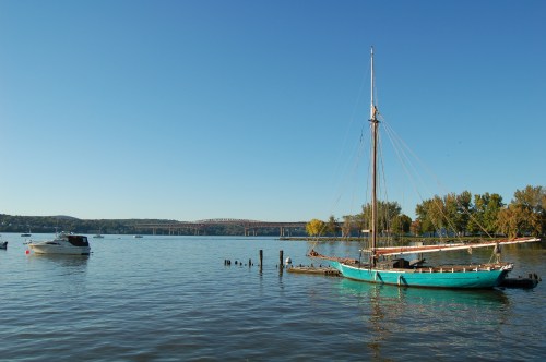 A calm river scene featuring a sailboat and a motorboat, with trees and a bridge in the background under a clear blue sky.