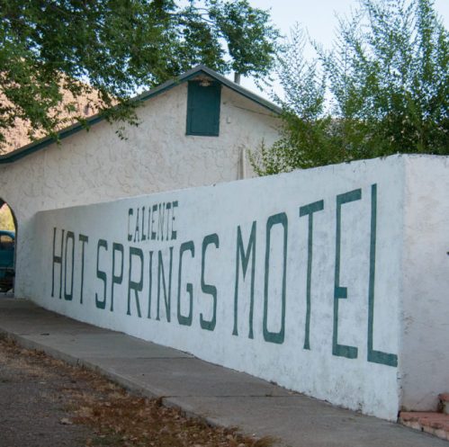 A white wall with green lettering reads "Caliente Hot Springs Motel," surrounded by trees and a building.