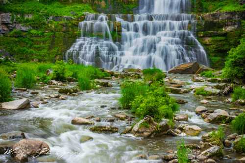 A serene waterfall cascades over rocks, surrounded by lush greenery and a gentle stream flowing in the foreground.