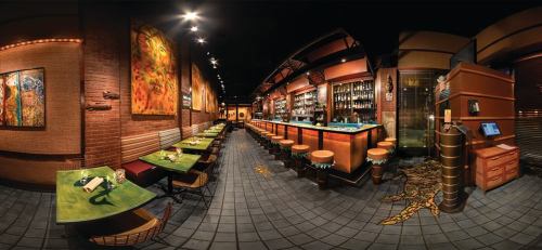 A panoramic view of a stylish bar with colorful artwork, wooden furniture, and a well-stocked bar area.