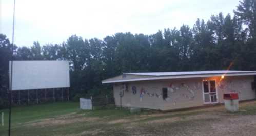 A small building with a porch and decorations, next to a large blank outdoor screen, surrounded by trees.