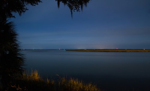 A serene night scene of a calm waterway with distant lights and grassy edges under a twilight sky.