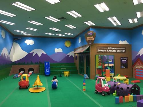 Colorful indoor play area with soft toys, slides, and a mural of mountains and a sun on the walls.
