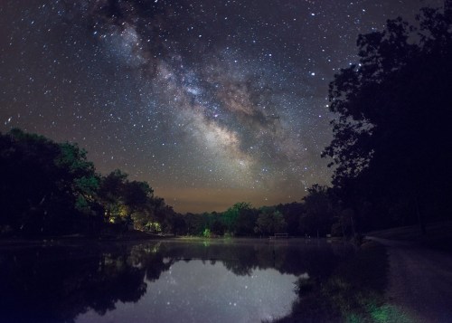 A serene night scene featuring a starry sky and the Milky Way reflected in a calm lake surrounded by trees.