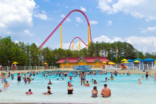 A busy water park scene with people swimming in a pool, colorful slides, and trees under a blue sky with clouds.