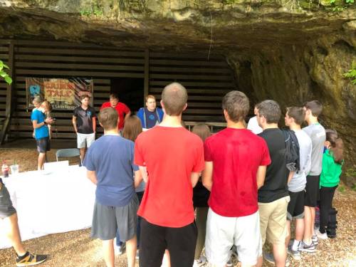 A group of people gathered outside a cave, listening to speakers at an event with a banner in the background.