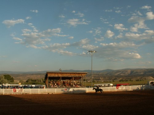 A rodeo arena with a crowd under a wooden structure, surrounded by mountains and a blue sky with scattered clouds.