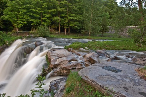 A serene waterfall cascades over rocks, surrounded by lush green trees and a peaceful landscape.