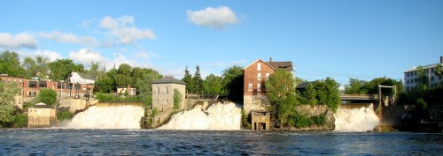A scenic view of a waterfall cascading over rocks, with buildings and greenery along the riverbank under a blue sky.