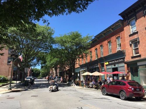 A sunny street scene with trees, cafes, and parked cars, showcasing a lively urban atmosphere.