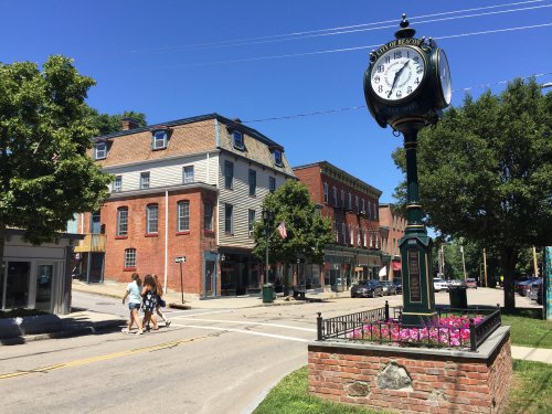 A historic town square featuring a clock tower, brick buildings, and pedestrians walking on a sunny day.