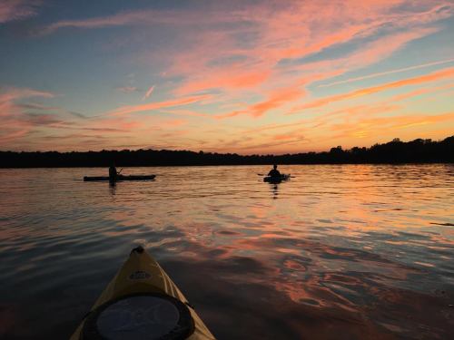 Two kayakers paddle on a calm lake at sunset, with vibrant pink and orange clouds reflecting on the water.