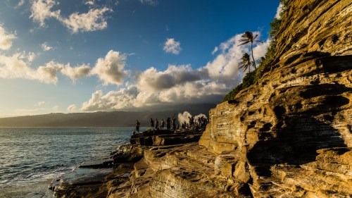 A rocky coastline with people exploring, palm trees, and a dramatic sky with clouds at sunset.