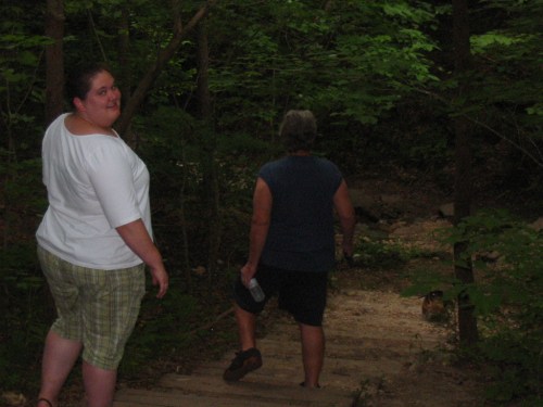 Two people walking down a wooded path, one looking back at the camera while the other walks ahead.
