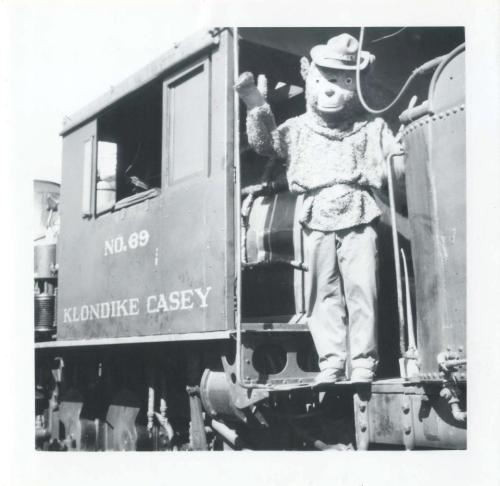A bear mascot in a hat waves from the window of a vintage train labeled "Klondike Casey."