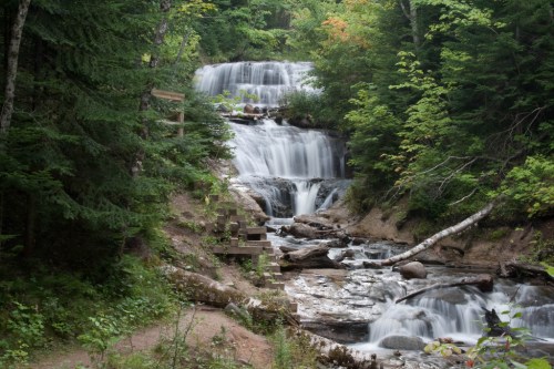 A serene waterfall cascades down rocky steps, surrounded by lush green trees and a peaceful forest setting.