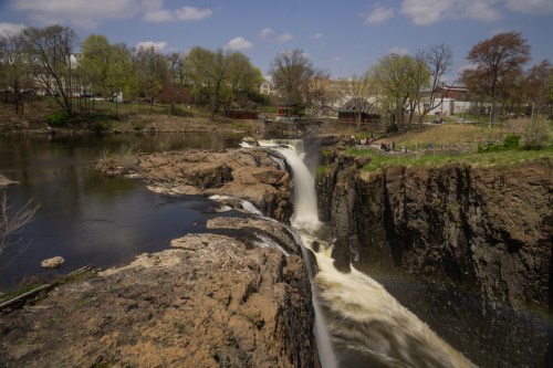 A scenic view of a waterfall cascading over rocky cliffs, surrounded by greenery and a calm river under a blue sky.